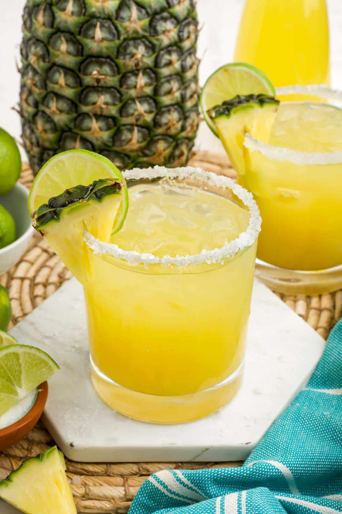 A pineapple margarita on a white coaster with fresh pineapple whole and cut on the table.