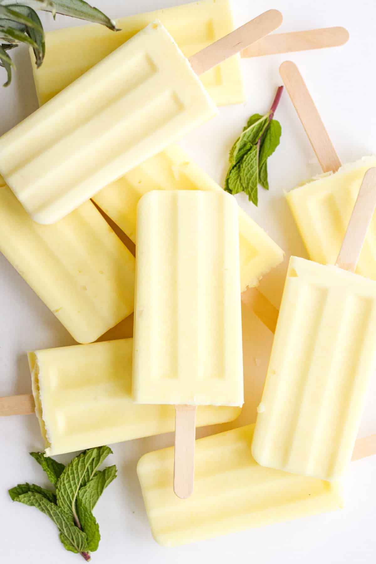 Dole whip popsicles laying in different directions on a white counter with some stacked and garnished with fresh mint.