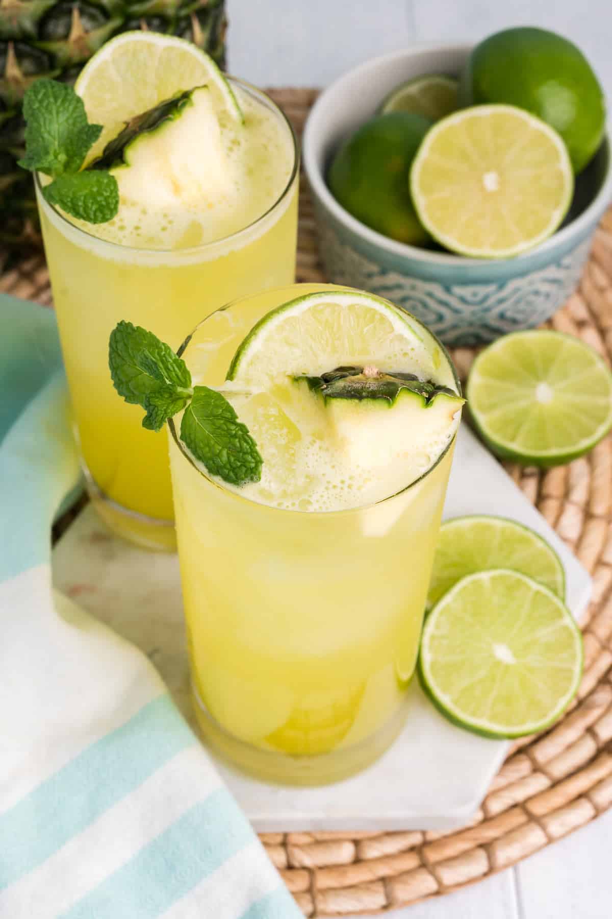 Glasses of pineapple agua fresca on the table with a bowl of limes and garnished with fresh pineapple and mint.