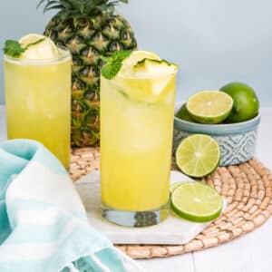 Glasses of pineapple agua fresca on the table with limes and a pineapple in the back.