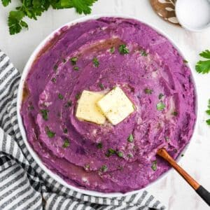 A bowl of purple mashed potatoes on the table with butter on top and a spoon in the bowl.