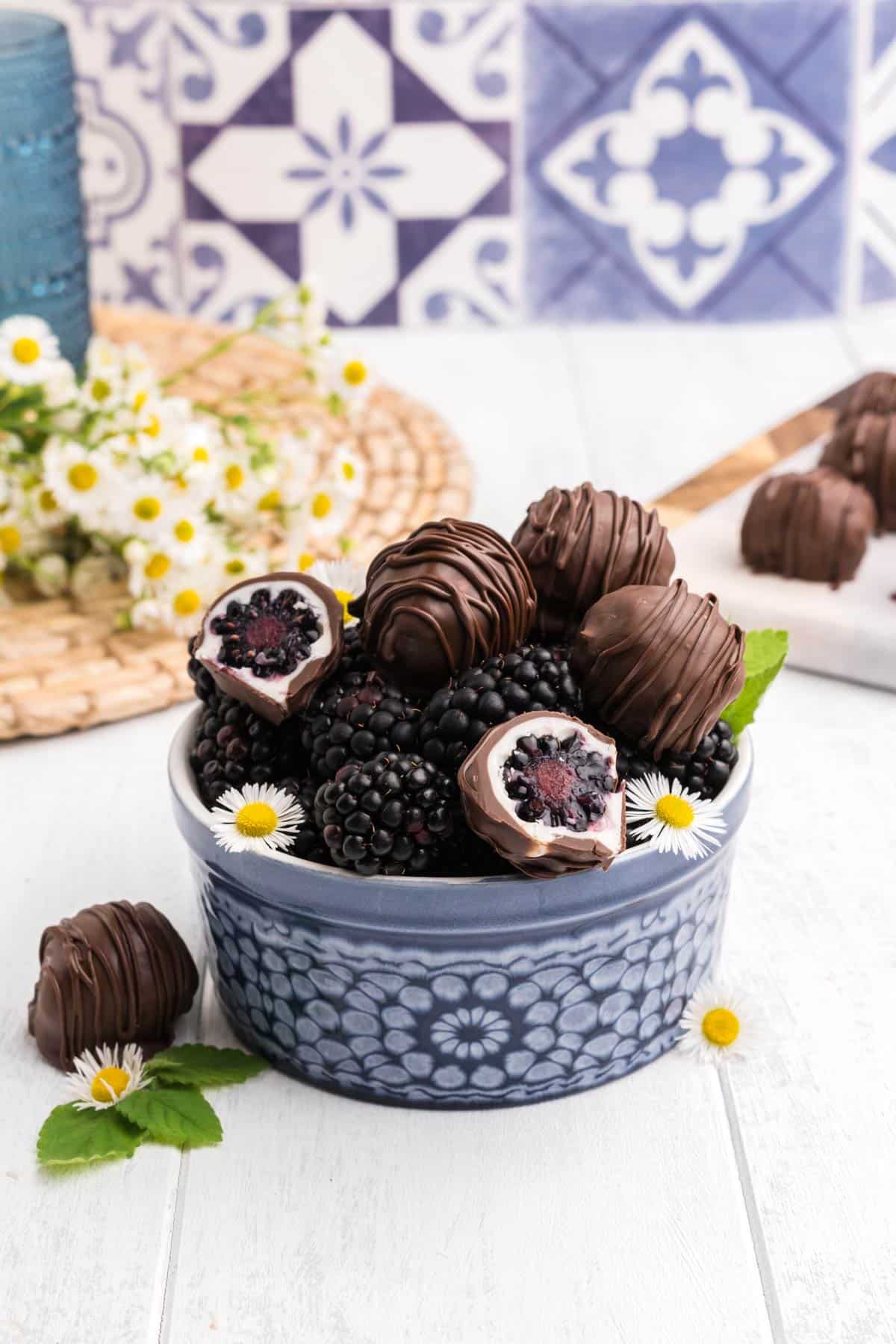 Chocolate covered blackberries in a blue bowl on the table with white and yellow flowers.