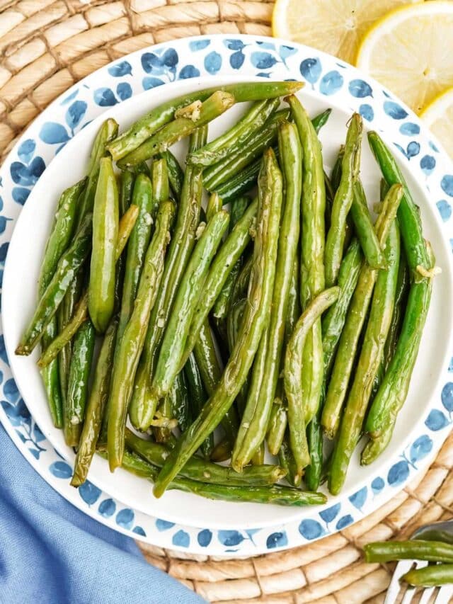 A plate of oven roasted green beans on the table with lemon slices and a blue napkin.