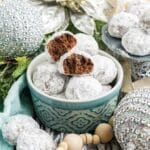 A bowl of chocolate snowball cookies on a table decorated with silver holiday balls and decorations.