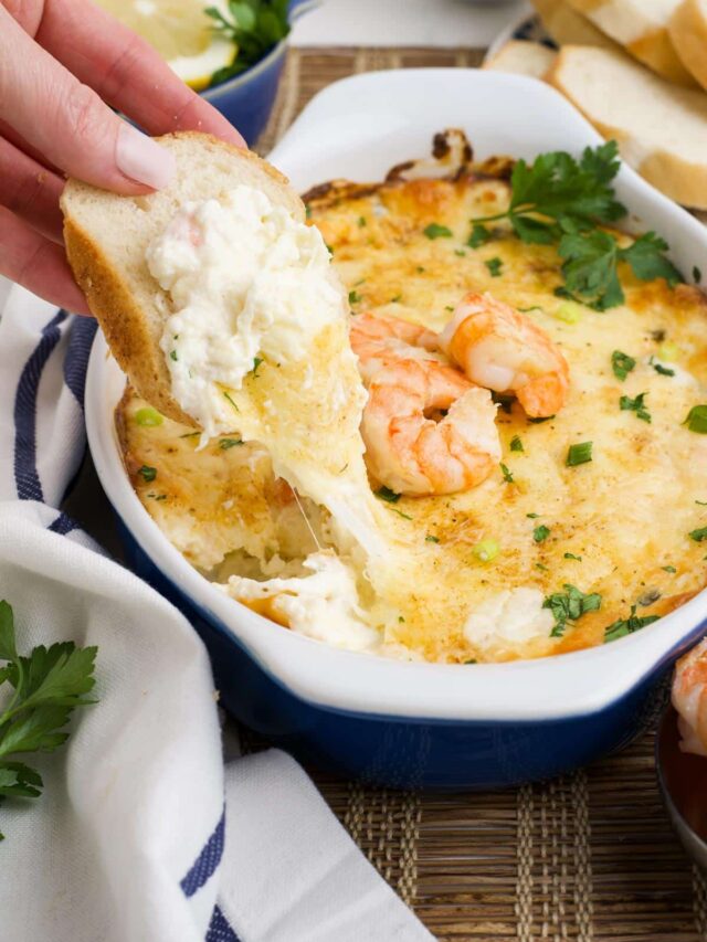A hand with a piece of bread dipping into the cheesy shrimp dip to show cheese pull.