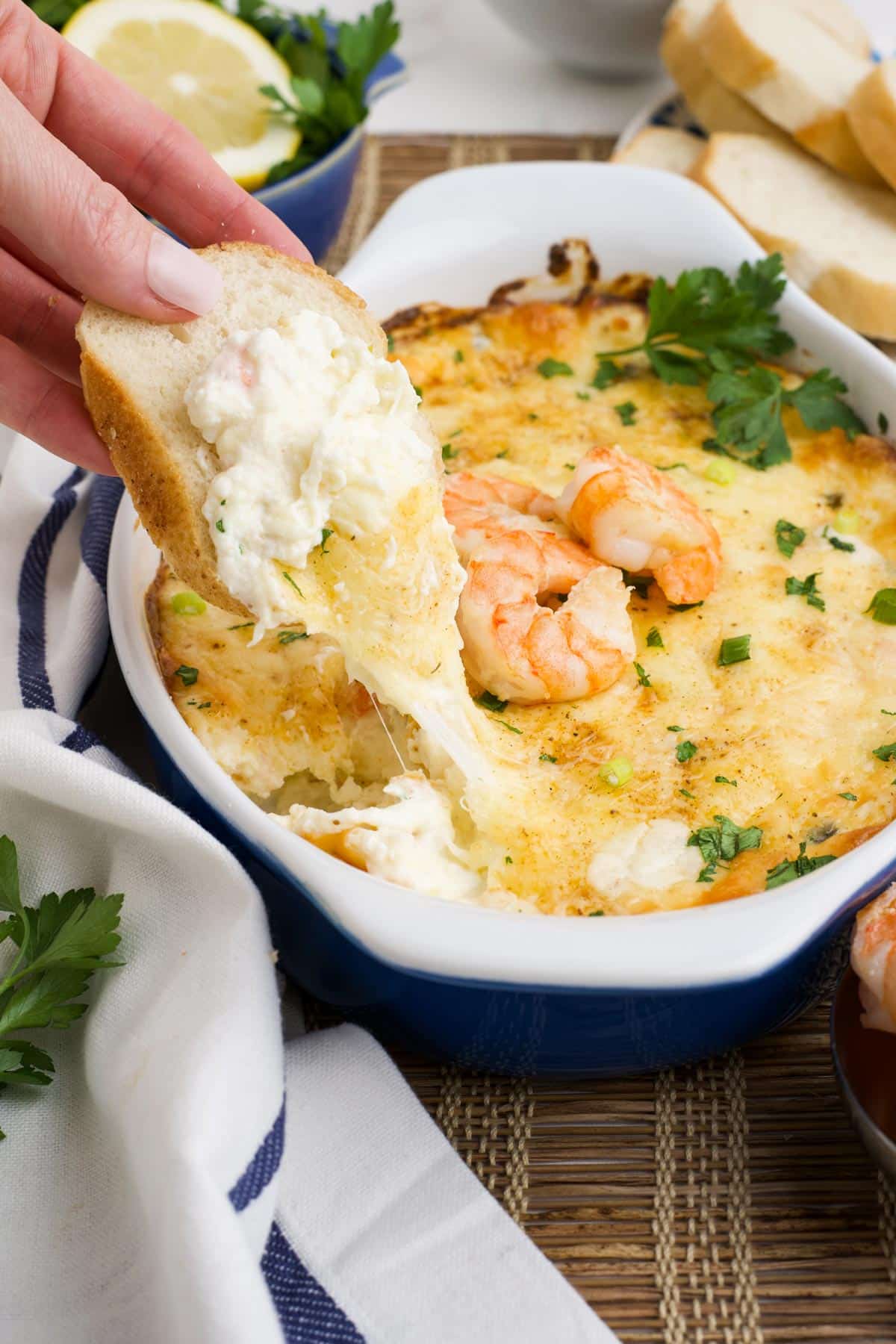 A hand with a piece of bread dipping into the cheesy shrimp dip to show cheese pull.