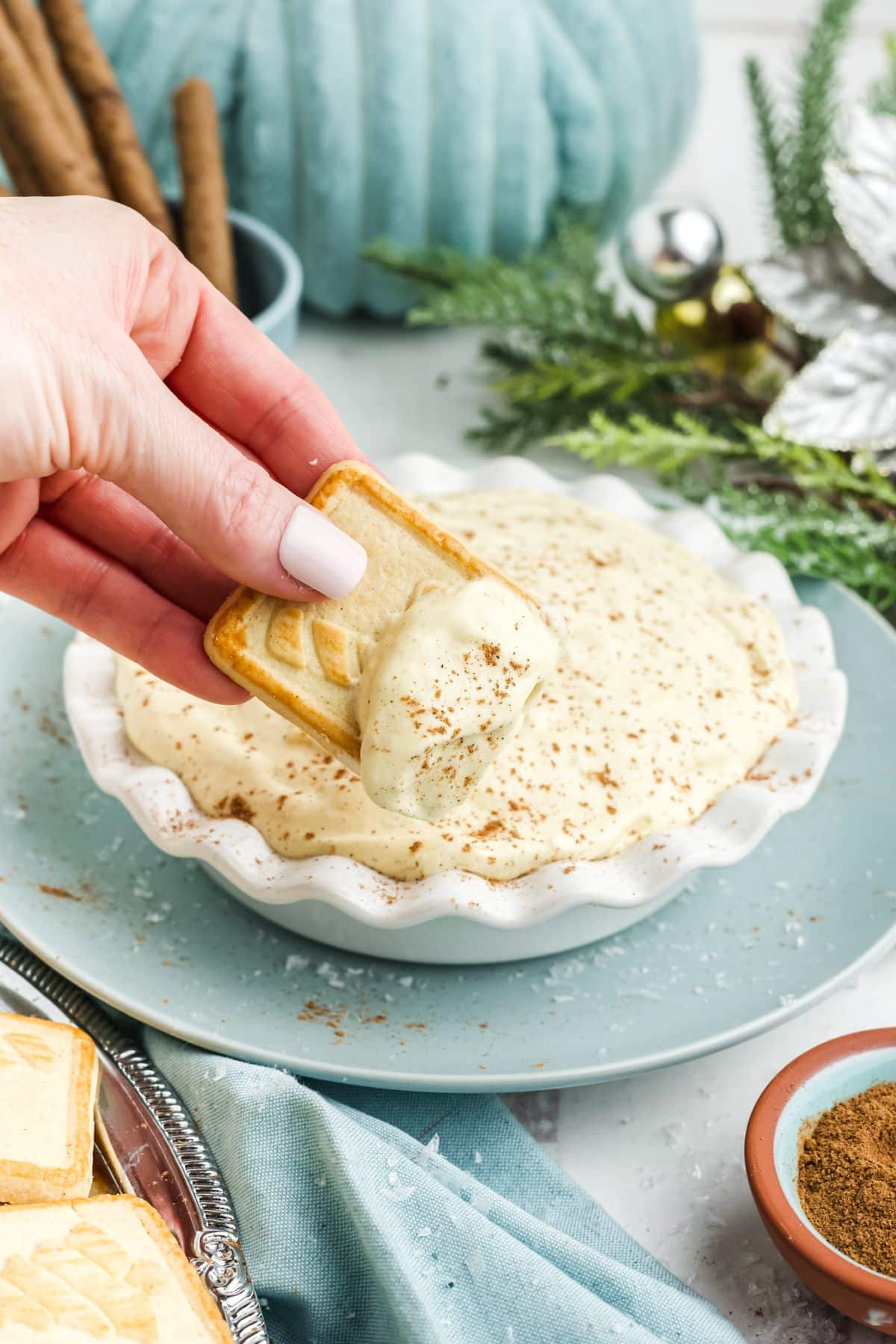 A hand dipping a cookie into a sweet eggnog dip in a bowl on a holiday table.