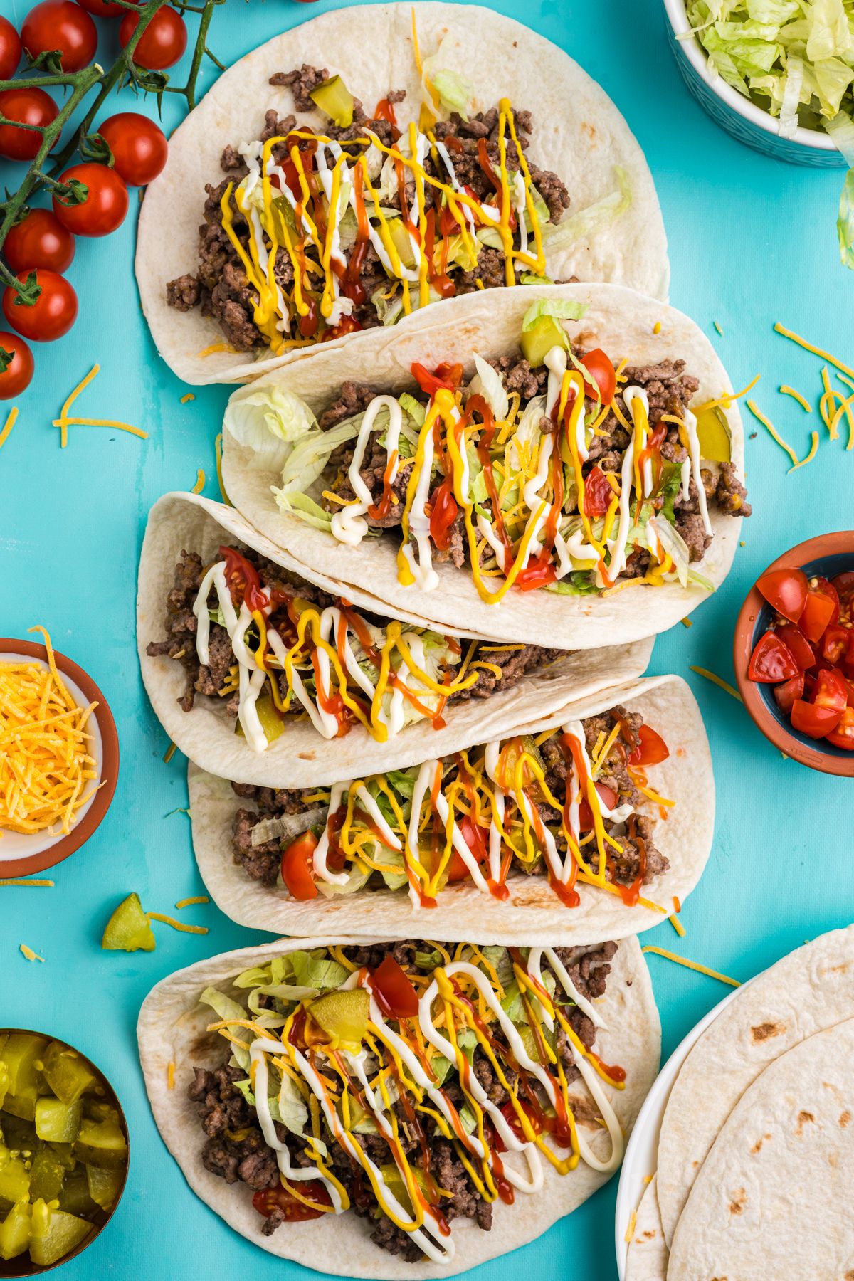 Cheeseburger tacos on the table with bowls of condiments surrounding them.