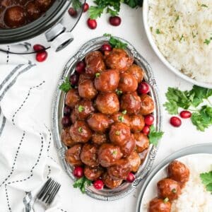 Cranberry meatballs on a oval silver platter with fresh cranberries and parsley around on the table.