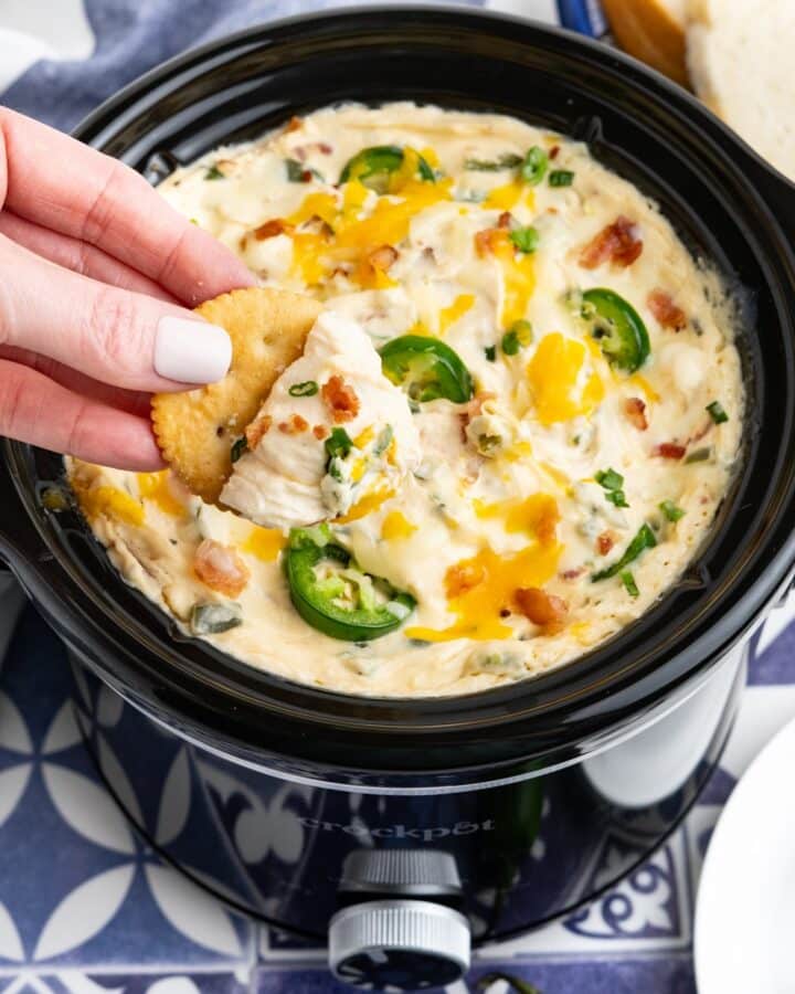 Crockpot jalapeno popper dip served up in a slow cooker with a hand dipping a cracker.