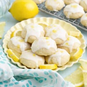 Lemon ricotta cookies on a yellow plate with more cookies on a wire rack in the background.