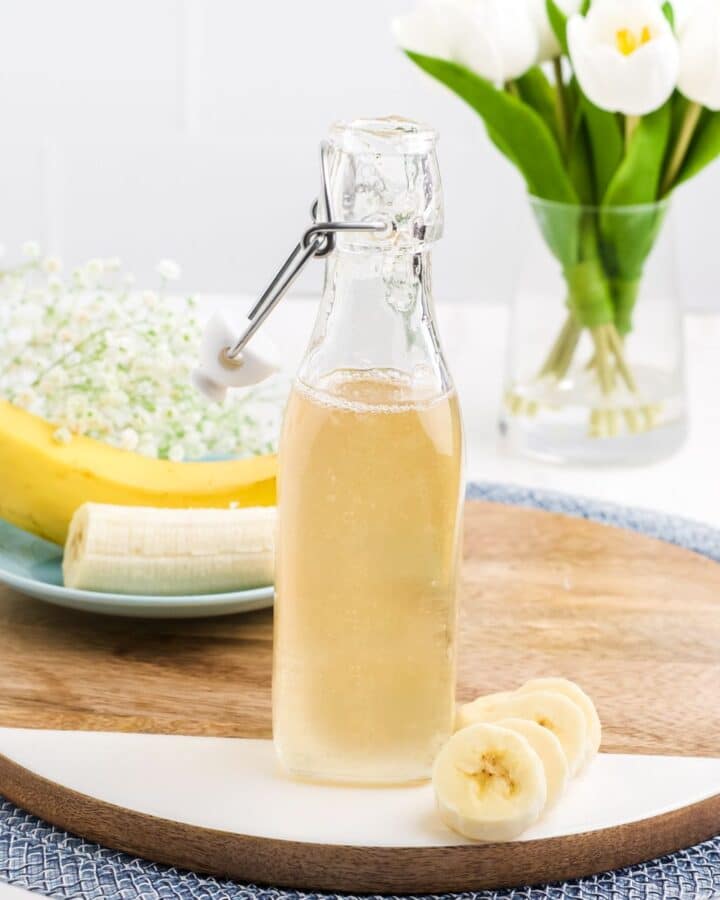A bottle of banana syrup on a wooden platter on the table with a whole banana and some slices in front.
