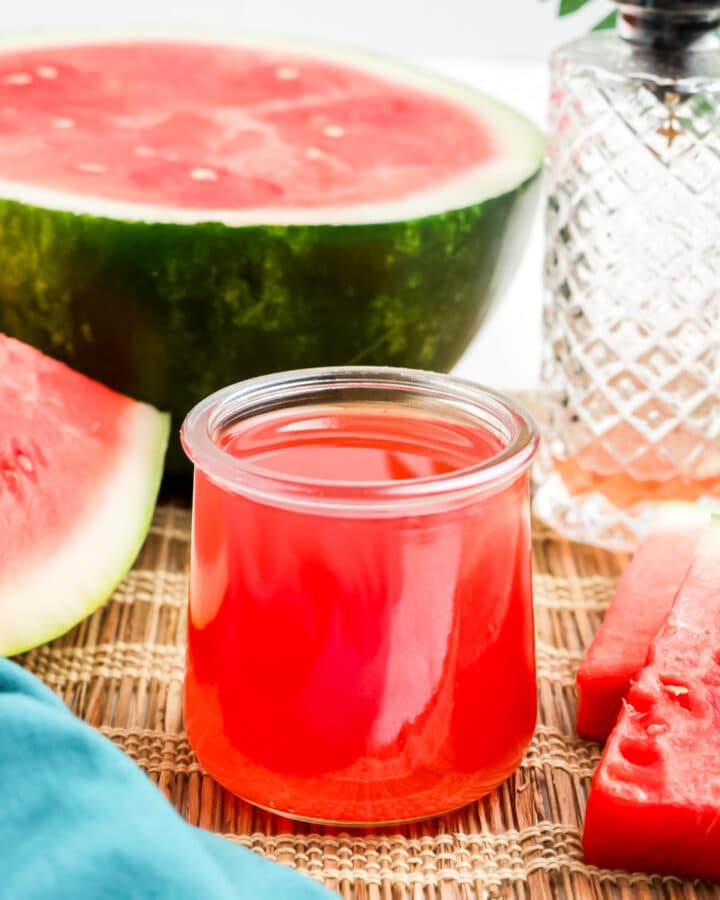 A little jar of watermelon simple syrup on the table with fresh watermelon in the back.