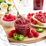 A jar of raspberry jalapeno jam on a wooden cutting board surrounded by berries and jalapeno slices while a spoon lifts up a bit from the jar.