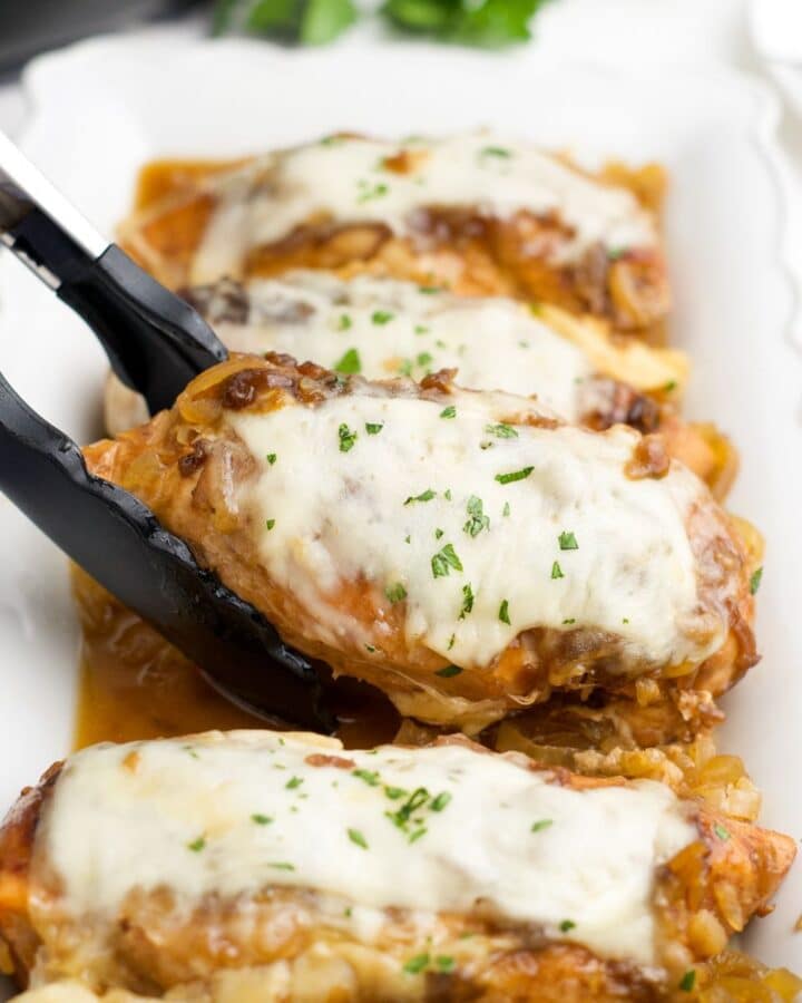 A pair of tongs lifting a piece of slow cooker french onion chicken from a plattter.