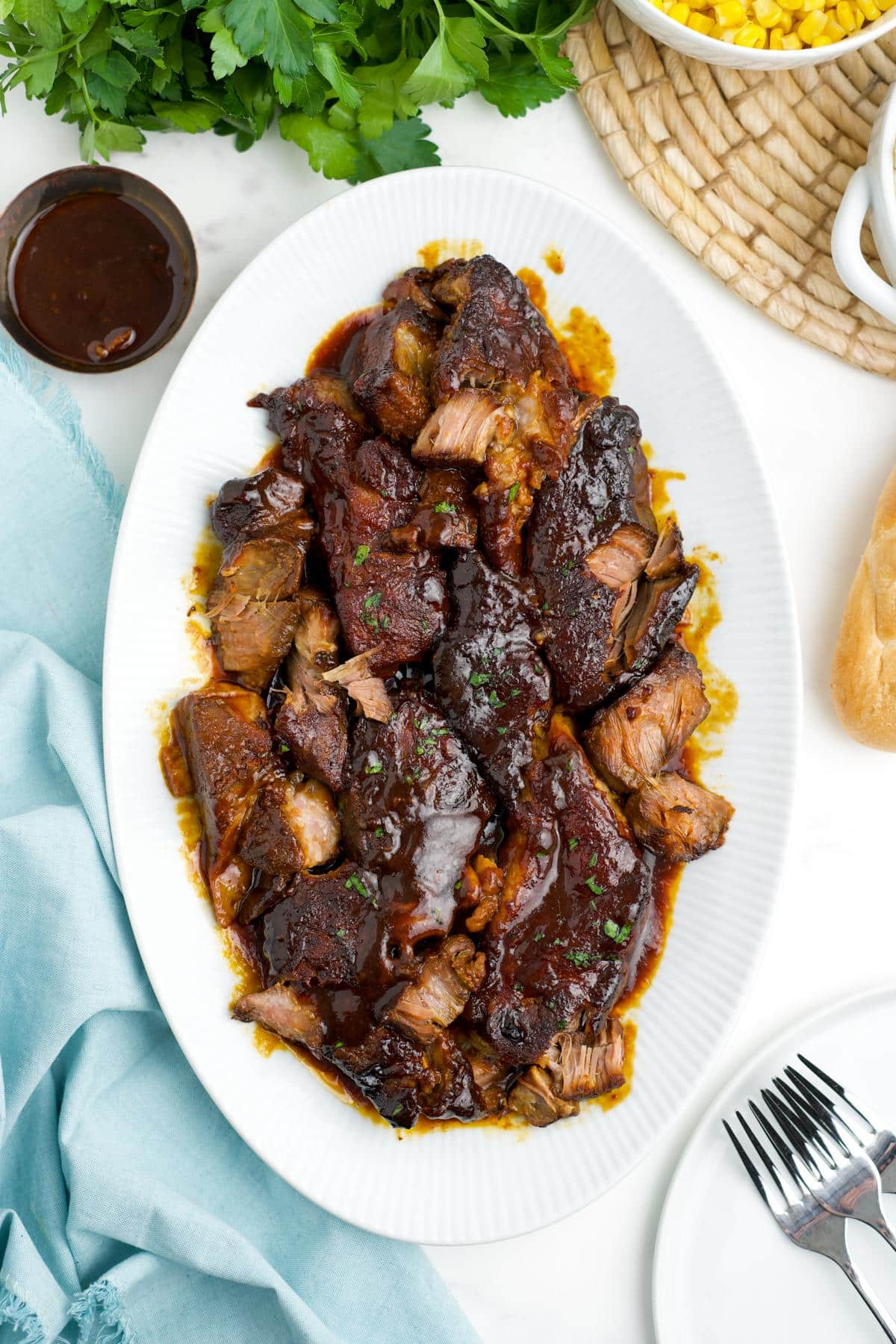 A platter of slow cooker boneless pork ribs on a table with a bowl of sauce and forks to the side.