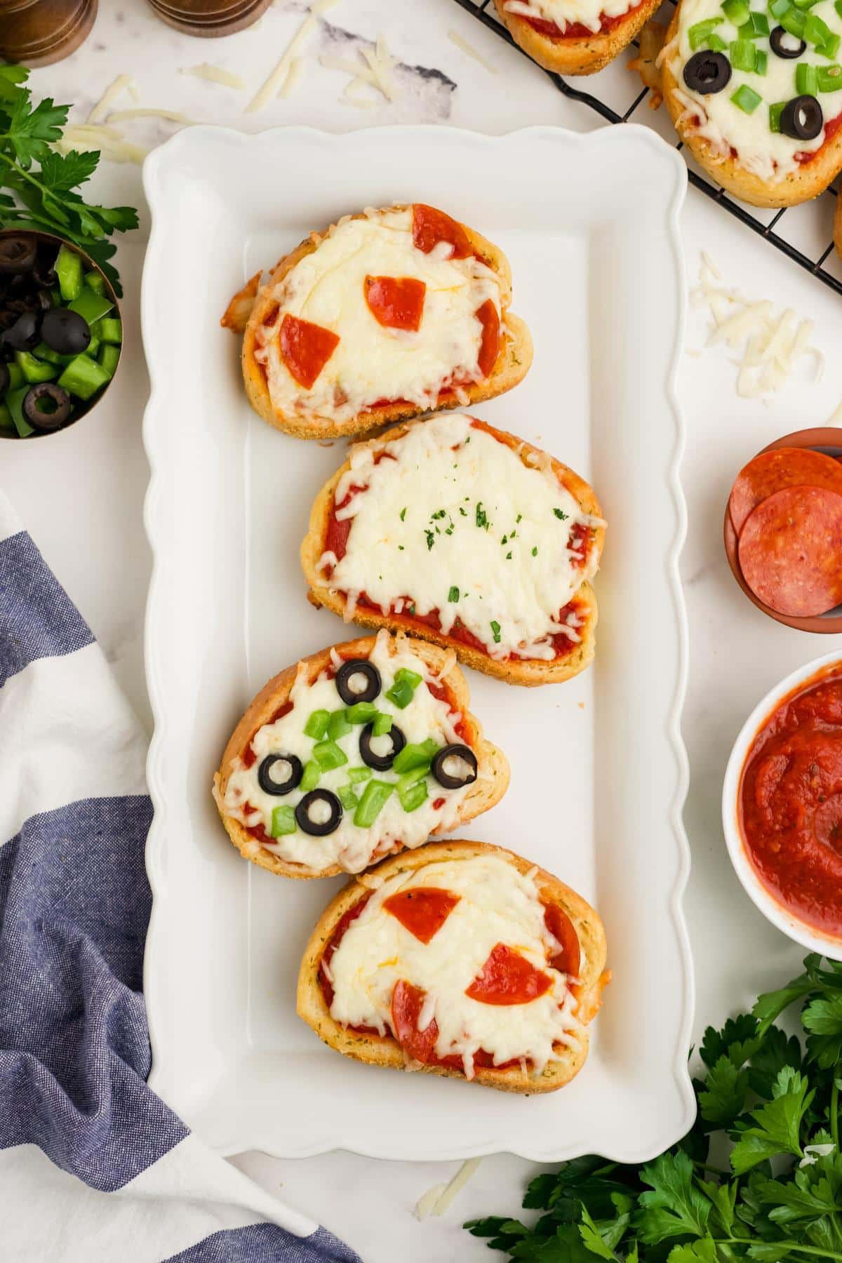 Four Texas toast pizzas on a white platter with bowls of ingredients to the side.