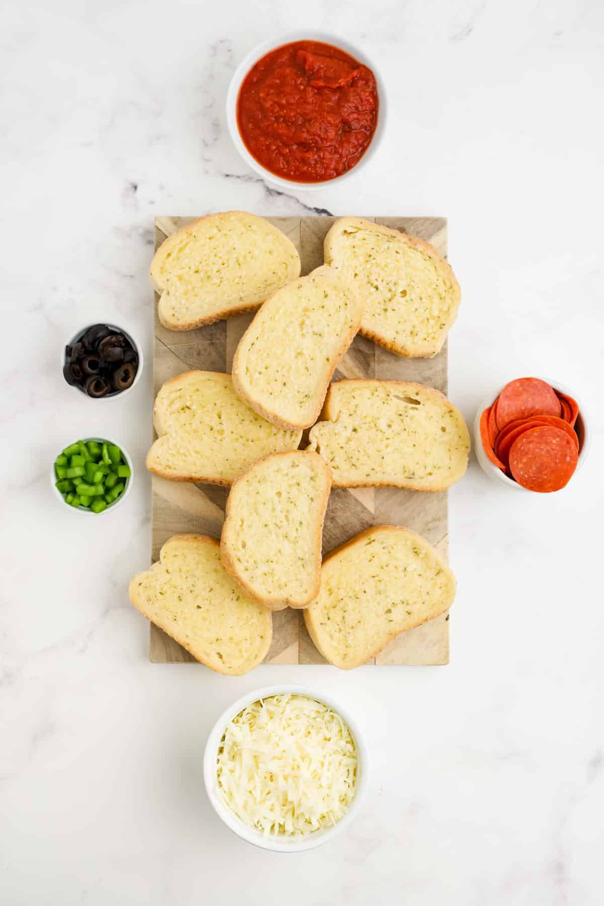 Ingredients to make pizza on Texas toast on the table in little bowls and the bread laid out on a wooden board.