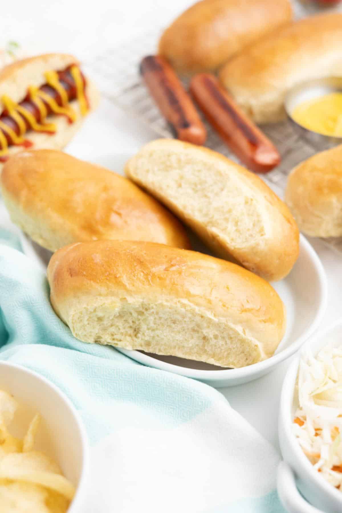 Homemade hot dog buns on a plate with ingredients to make hot dogs surrounding them.