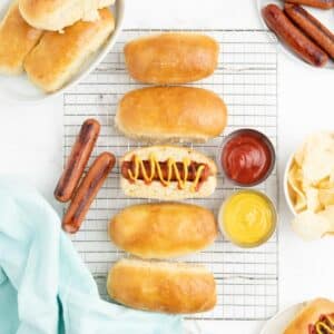 Homemade hot dog buns on a wire rack with one in the middle filled with a hot dog topped with mustard and ketchup.