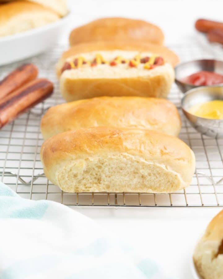 Homemade hot dog buns on a table with condiments and grilled hot dogs.