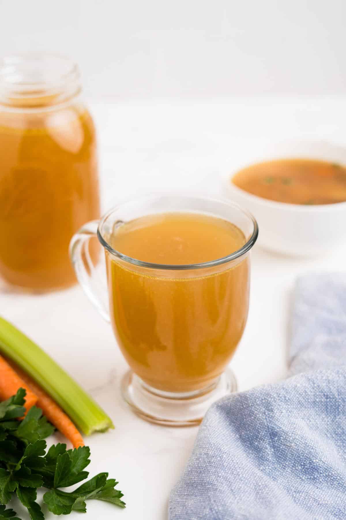 Slow cooker chicken broth in a cup in front of a jar and bowl of more.