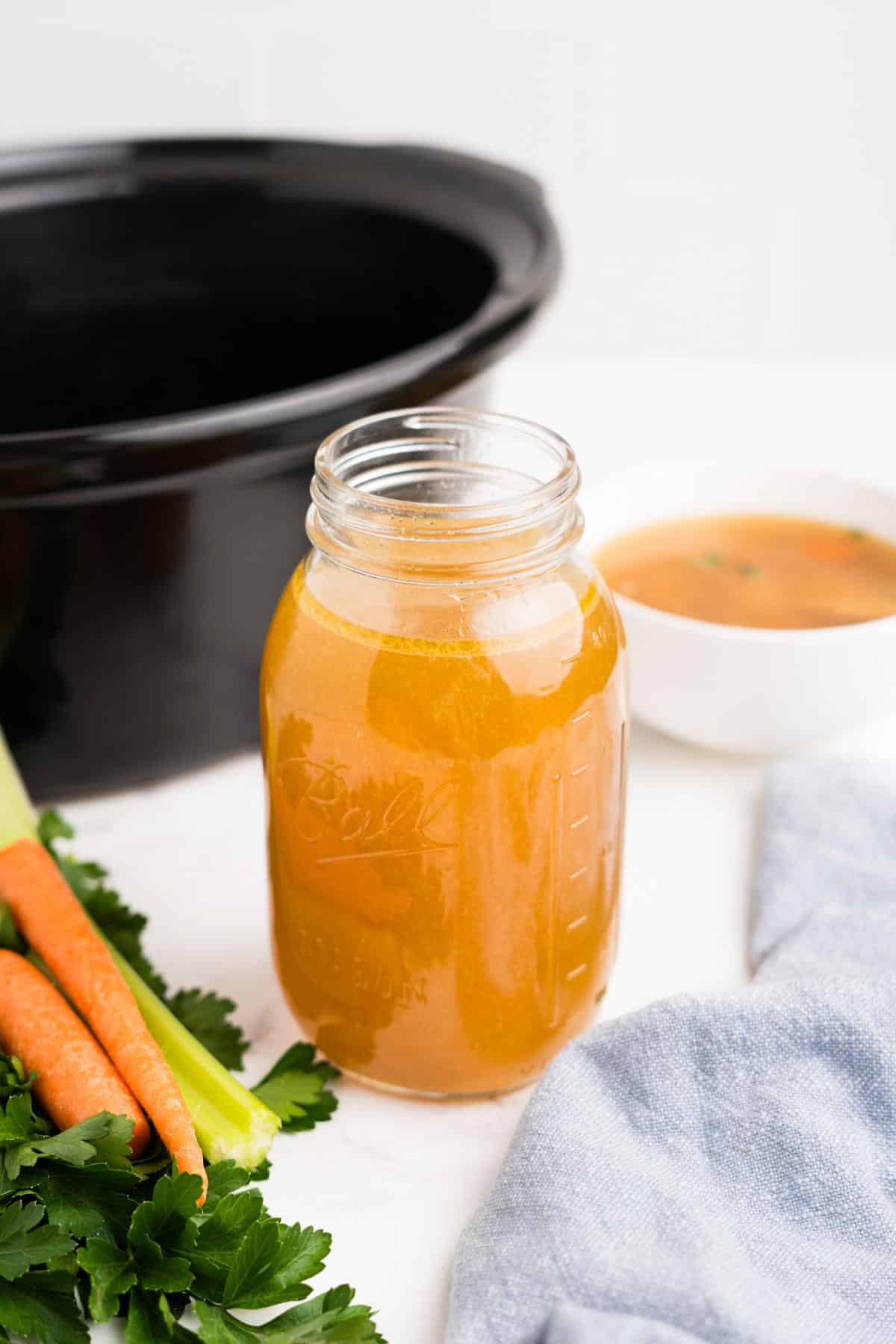 A jar of slow cooker rotisserie chicken broth in front of the pot and next to some vegetables.