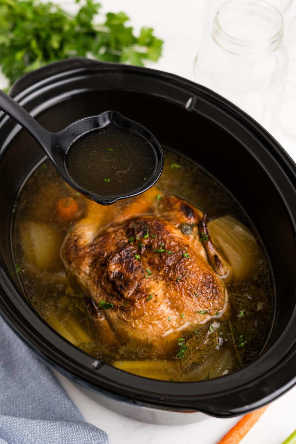 A ladleful of chicken broth held up over a slow cooker.
