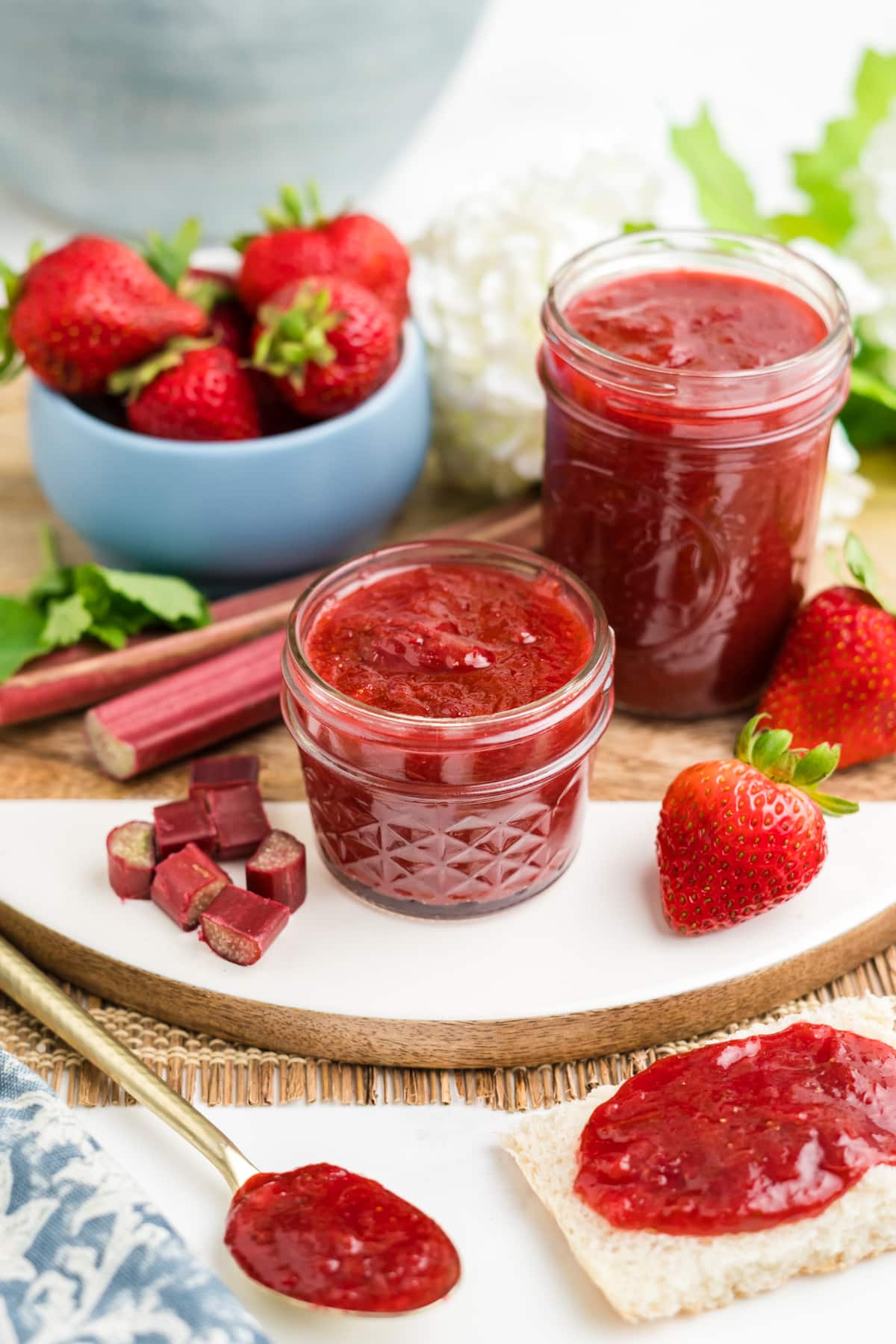 An image of strawberry and rhubarb preserves with ingredients surrounding it and a spoon and jam covered bread in the front.