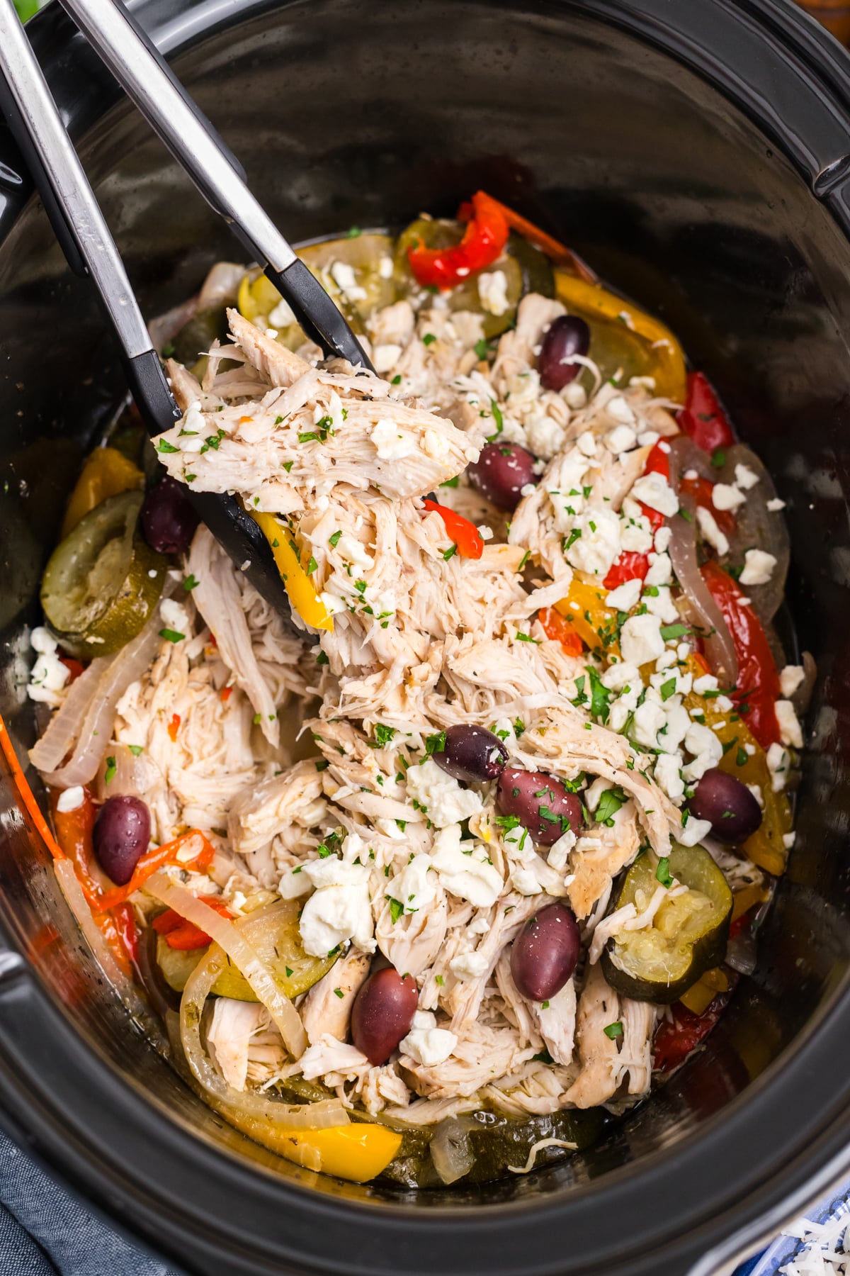 Cooked Greek chicken in a slow cooker with tongs lifting up some of the chicken. 