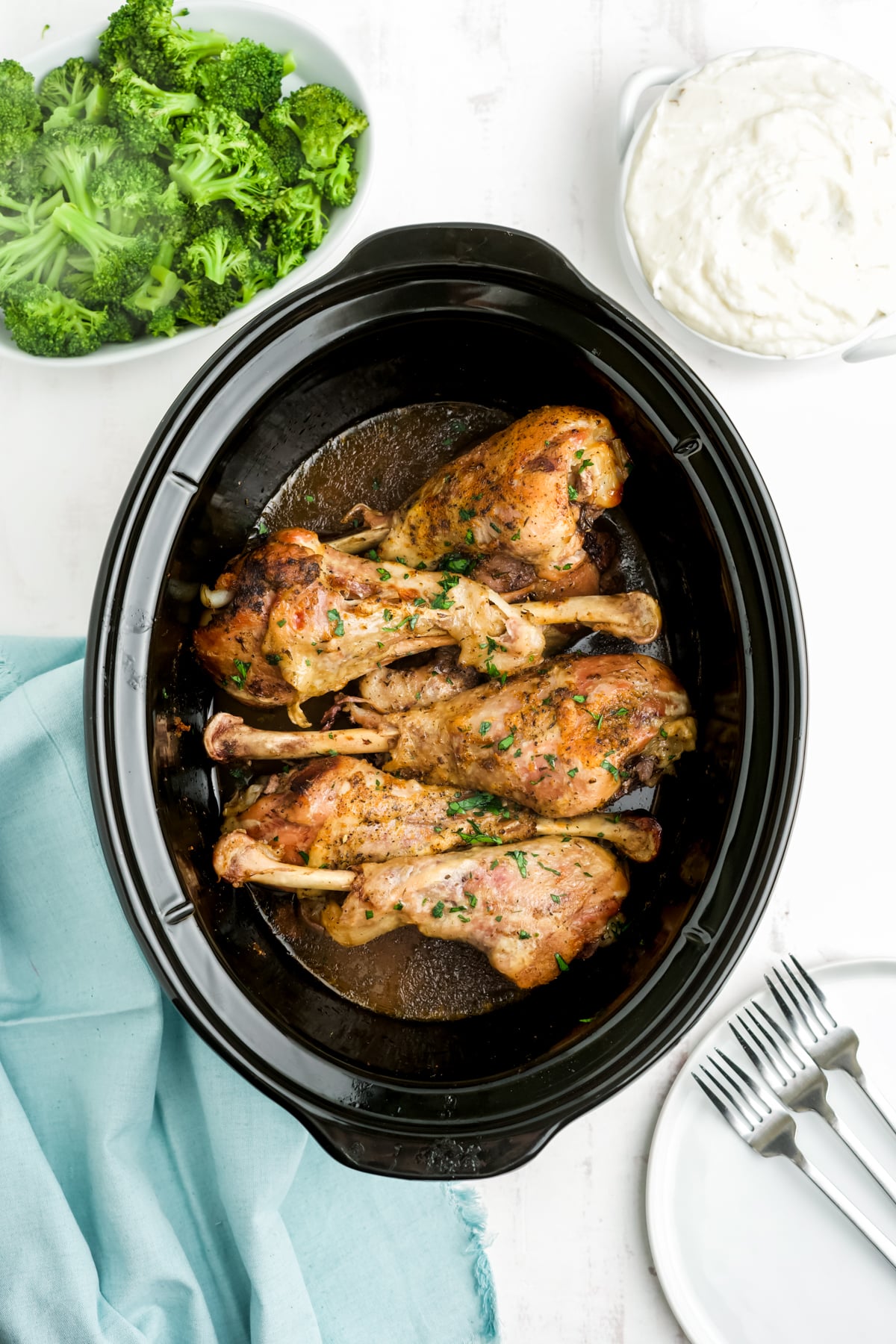 Slow cooked turkey legs in the slow cooker with bowls of broccoli and mashed potatoes to the side. 