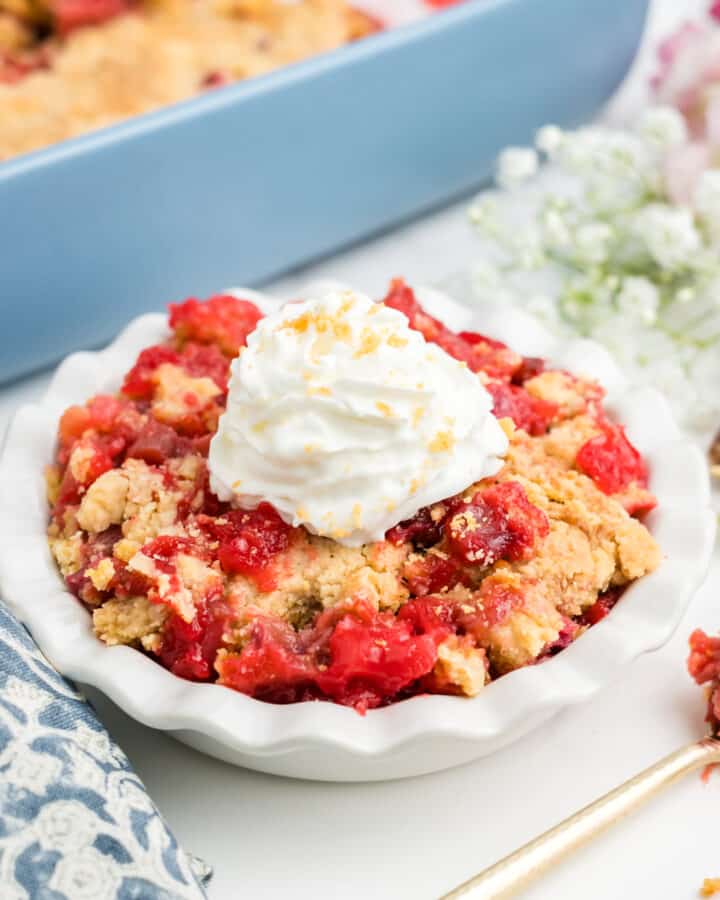 A dish of rhubarb dump cake topped with whipped cream in front of the pan of more.