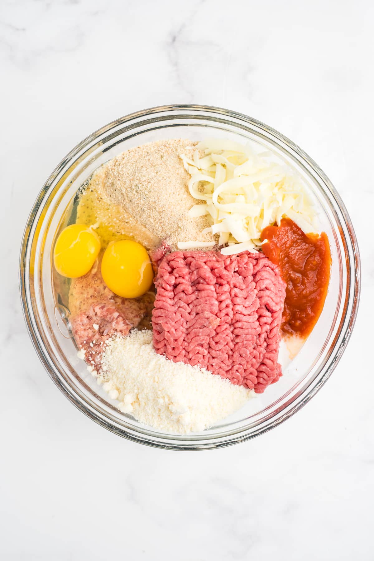Ground beef, sausage, breadcrumbs, eggs, milk, Parmesan, mozzarella, seasonings, sauteed onion, garlic, and reduced marinara in a bowl.
